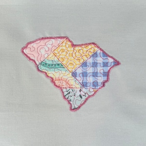 May include: A patchwork quilt in the shape of the state of South Carolina. The quilt is made of different colored fabrics, including blue, yellow, pink, and green. The quilt is stitched together with pink thread.
