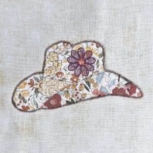 Può includere: Applicazione di cappello da cowboy in tessuto con un motivo floreale nei toni del giallo, marrone e rosa. Il cappello presenta un grande fiore viola ricamato al centro ed è delineato da cuciture marroni. Lo sfondo è un tessuto beige chiaro.
