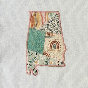 May include: A patchwork quilt in the shape of the state of Alabama. The quilt is made of various fabrics with floral and geometric patterns in shades of green, brown, pink, and white.