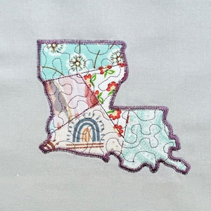 Può includere: Una trapunta patchwork a forma dello stato della Louisiana. La trapunta è realizzata con diversi tessuti con motivi floreali e geometrici in tonalità di blu, rosa, rosso e bianco. La trapunta è bordata con una cucitura viola.