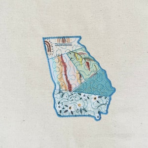 Può includere: Un'applique in tessuto patchwork dello stato della Georgia. L'applique è realizzata con diversi tessuti blu, verdi e marroni con motivi floreali. L'applique è contornata da filo blu.