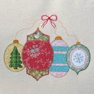 Pode incluir: Enfeites de Natal aplicados em várias formas e padrões, incluindo uma árvore de Natal, desenhos florais e um floco de neve. Os enfeites são em tons de vermelho, verde, azul e rosa, conectados por fios dourados e um laço vermelho. O fundo é um tecido neutro.
