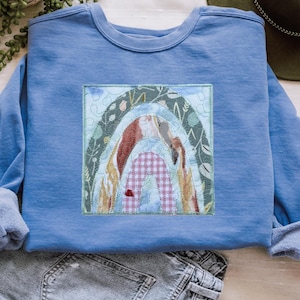 May include: A light blue sweatshirt featuring a colorful rainbow applique. The rainbow design incorporates various patterns and colors, including a gingham pattern. The applique is framed by a square border with decorative stitching. The sweatshirt is made of a soft material.