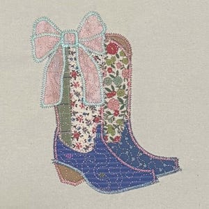 Puede incluir: Botas de vaquero aplicadas con un lazo rosa. Las botas presentan tela floral y estampada en tonos de azul, rosa y verde. El lazo tiene un ribete brillante. Las botas tienen tacones marrones y están cosidas sobre un fondo neutro.