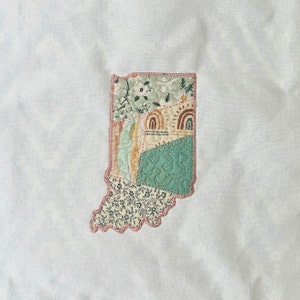 Può includere: Una trapunta patchwork a forma dello stato dell'Indiana. La trapunta è realizzata con diversi tessuti in tonalità di verde, marrone e bianco. La trapunta è cucita insieme con un punto decorativo.