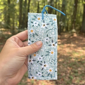 Op de afbeelding: Een rechthoekige, gewatteerde boekenlegger met een lichtblauw bloemenpatroon met witte madeliefjes en gele centra. De boekenlegger heeft een blauwe rand en een bijpassende blauwe koordlus aan de bovenkant.