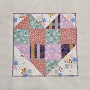 Op de afbeelding: Een patchwork hartontwerp op een vierkant stuk stof. Het hart is samengesteld uit verschillende stoffen met patronen in roze, groen, paars en wit, aan elkaar gestikt. De randen zijn afgewerkt met een paarse zigzagsteek.