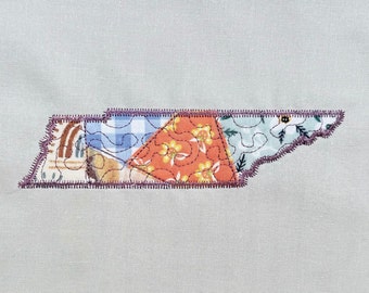 May include: A patchwork fabric applique of the state of Tennessee. The applique is made of various fabrics with floral and geometric patterns in shades of blue, orange, green, and brown. The applique is outlined with a purple thread.