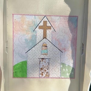 Puede incluir: Una pieza de bordado cuadrada sobre tela que muestra el diseño de una iglesia con una cruz color canela, una puerta con estampado floral y una pequeña ventana arqueada. El fondo es tela clara estilo acuarela con costuras y colinas verdes.