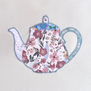 May include: A fabric teapot with a floral design. The teapot is white with pink and burgundy flowers, green leaves, and a blue geometric pattern on the lid. The handle and spout are light blue, and the edges are outlined with gray stitching.