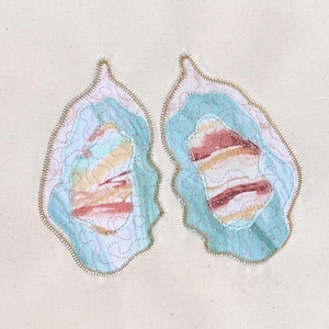 Può includere: Due opere d'arte tessile astratte, ciascuna con un design a strati nei toni del celeste, rosa e giallo. I pezzi hanno un bordo smerlato e cuciture dorate, su uno sfondo color crema. Fatto a mano.