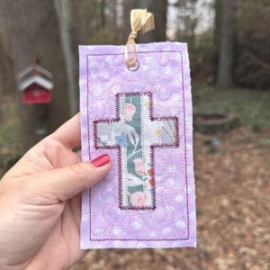 May include: A rectangular fabric bookmark in light purple with a white swirling pattern and a central cross applique. The cross features a green background with a floral print of pink tulips and blue leaves, finished with dark purple stitching.