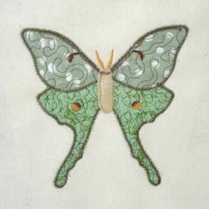 Op de afbeelding: Een stoffen vlinderapplicatie met groen en wit patroonvleugels en een beige lichaam. De vleugels hebben een gedetailleerde, gestikte omtrek en hebben een mix van effen groene en witte wervelpatronen. De vlinder is gecentreerd op een crèmekleurige stof.