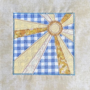 Può includere: Un blocco di tessuto quadrato con un disegno a raggiera. I raggi del sole sono gialli e dorati, che irradiano da un cerchio centrale. Lo sfondo è un motivo a quadretti blu e bianco, con un bordo beige chiaro.