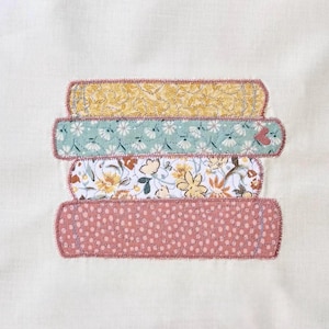 Puede incluir: Una pila de cuatro libros de tela con patrones florales y de lunares decorativos. Los libros son en tonos de amarillo, verde azulado, blanco y rosa, con un borde cosido. Un pequeño corazón es visible en el libro verde azulado.