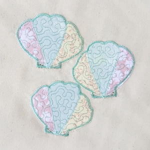 Può includere: Tre toppe di tessuto a forma di conchiglia. Ogni toppa è divisa in tre sezioni: rosa floreale, azzurro con cuciture ondulate e giallo floreale. I bordi sono delineati con cuciture verdi, su uno sfondo beige chiaro.