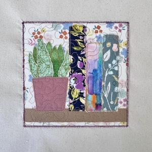 May include: A fabric art piece featuring a potted succulent and three stylized books on a shelf. The books have floral and watercolor patterns. The background is a floral print, and the piece is framed with a purple stitched border.