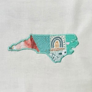 May include: A patchwork fabric appliqué of the state of North Carolina in teal, pink, and brown. The appliqué is stitched onto a white fabric background.
