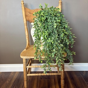 May include: A distressed wooden chair with a carved back supports a white planter filled with trailing green leaves. The chair has a light brown finish, set against a grey backdrop. The image evokes a rustic, natural feel.