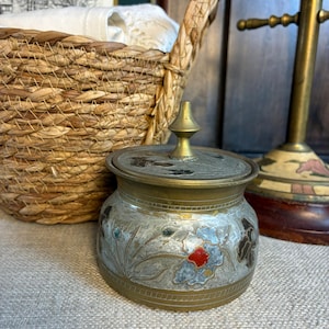 May include: A small, round brass container with a removable lid featuring a pointed finial. The exterior surface is decorated with etched floral patterns in muted tones of blue, red, and dark brown against a weathered, metallic silver-coloured finish.