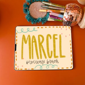 May include: A digital tablet displaying a graphic design of the word "MARCEL" in a yellow font with a textured effect. The text "procreate brush" is written below the word "MARCEL" in a smaller font. The tablet is on a red surface.