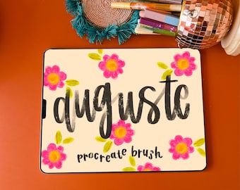 Może przedstawiać: Tablet z napisem "auguste" w czarnym skrypcie, otoczony różowymi kwiatami i zielonymi liśćmi. Słowa "procreate brush" znajdują się poniżej. Tablet leży na pomarańczowej powierzchni, wraz z uchwytem na długopisy w kształcie kuli dyskotekowej i długopisami.
