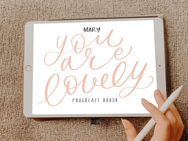 Pu&ograve; includere: Un tablet digitale che mostra un design di calligrafia rosa che dice "you are lovely" con il testo "Procreate Brush" sotto. Il tablet &egrave; su una superficie marrone testurizzata.