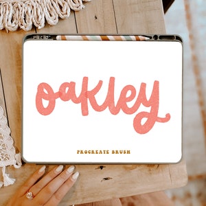 Puede incluir: Un dibujo digital del nombre "Oakley" en una fuente rosa manuscrita sobre un fondo blanco. El dibujo se muestra en la pantalla de una tableta con un lápiz óptico apoyado en la pantalla. El texto "Procreate Brush" se muestra debajo del nombre.