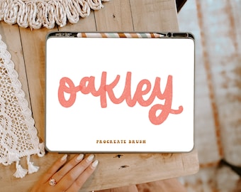 Può includere: Un disegno digitale del nome "Oakley" in un font rosa scritto a mano su uno sfondo bianco. Il disegno è visualizzato sullo schermo di un tablet con uno stilo appoggiato sullo schermo. Il testo "Procreate Brush" è visualizzato sotto il nome.