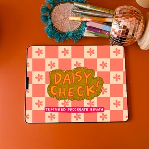 May include: A digital tablet with a daisy-themed design. The screen displays the words "DAISY CHECK!" in a yellow and orange font, with the text "TEXTURED PROCREATE BRUSH" below. The background is a pink and peach checkerboard pattern.