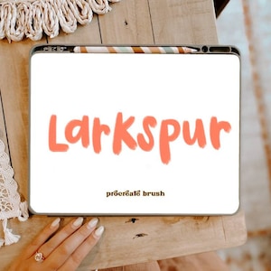 May include: A white digital tablet with a pink brush font that says "Larkspur" and the text "procreate brush" in brown font.