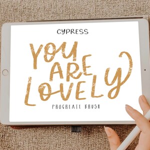 Pu&ograve; includere: Uno schermo di tablet digitale che mostra il testo "You are lovely" in un carattere dorato testurizzato. Il testo &egrave; scritto in uno stile informale, a mano libera. Le parole "Procreate Brush" sono scritte in un carattere pi&ugrave; piccolo sotto il testo principale.