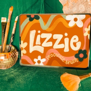 Puede incluir: Una tableta naranja con el nombre "Lizzie" y las palabras "procreate brush" en escritura blanca. Pinceles en un soporte de mosaico dorado y un pincel de maquillaje visible, todo sobre una superficie verde.