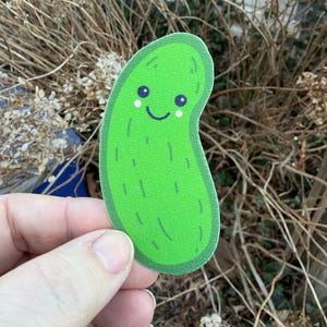 May include: A green, smiling pickle sticker with a cute face. The sticker has a textured surface and is held in front of a background of dried plants and foliage.