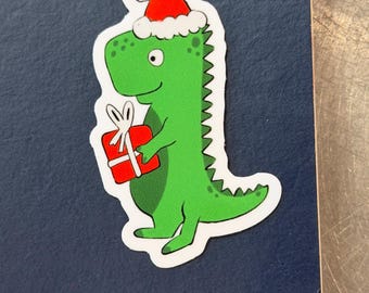 May include: A green dinosaur sticker wearing a red Santa hat and holding a red gift box with a white bow. The dinosaur has a white outline and is on a dark blue background.