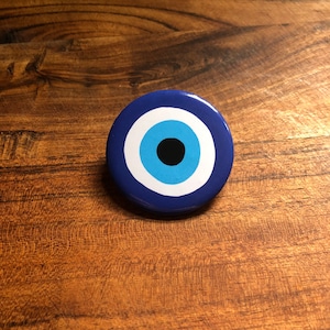 May include: A blue and white evil eye charm with a black pupil. The charm is round and has a glossy finish.