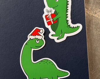 May include: Two green dinosaur stickers wearing Santa hats. One dinosaur is holding a red gift with a white bow. The stickers are on a dark blue surface. The stickers are white-bordered.