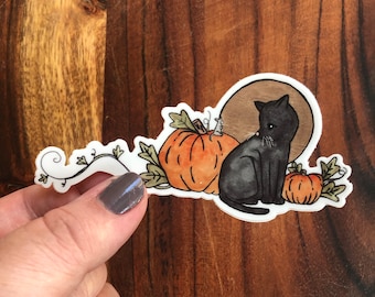 May include: A black cat sits next to two orange pumpkins with green vines and leaves. The cat is looking to the left, and the pumpkins are in the background. The cat is in the foreground. The background is a brown wooden surface.