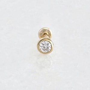 May include: A small, gold-colored stud earring with a round, clear gemstone. The gemstone is set in a bezel setting, and the earring post is visible. The earring is on a white marble surface.
