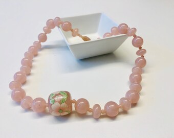 May include: A pink beaded necklace with a single floral patterned bead. The necklace is made of round, smooth beads and is displayed on a white surface.