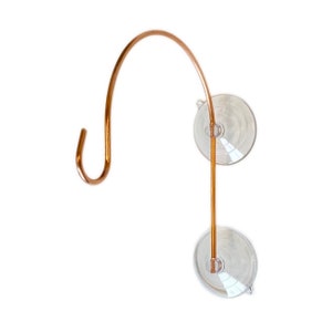 May include: A copper-colored, curved hook with a suction cup base. The hook is designed to hang items and attaches to smooth surfaces. The suction cups are clear and round, providing a secure hold. The hook is made of a durable metal.