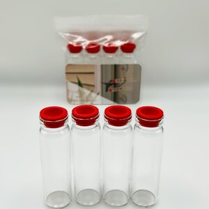May include: Four clear glass vials with bright red silicone stoppers arranged in a row. Behind them, a clear plastic bag contains additional identical vials, and a small rectangular card with a printed graphic is positioned in the center.
