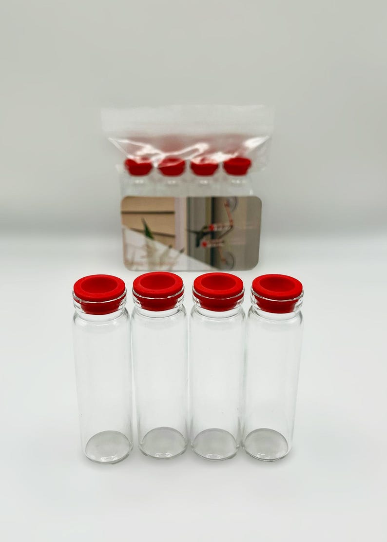 May include: Four clear glass vials with bright red silicone stoppers arranged in a row. Behind them, a clear plastic bag contains additional identical vials, and a small rectangular card with a printed graphic is positioned in the center.