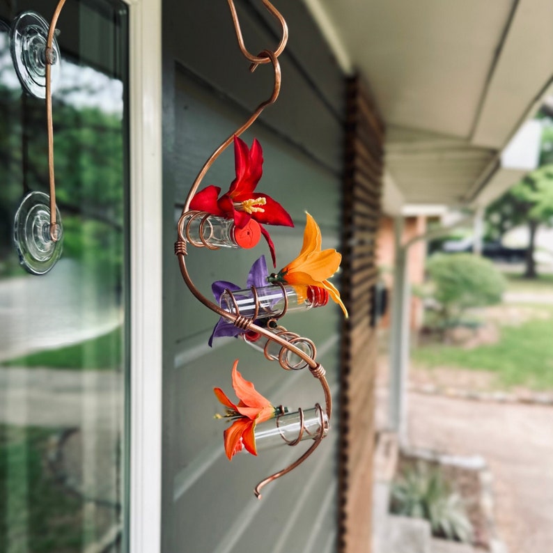 May include: A copper-colored, hanging hummingbird feeder with three glass vials, each holding a colorful artificial flower. The feeder is attached to a window with suction cups. The flowers are red, orange, purple, and yellow.