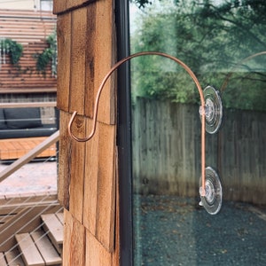 May include: A copper-colored, curved metal hook with a suction cup attachment. The hook is designed to hang items on a window or smooth surface. The hook is attached to the glass with two clear suction cups.