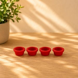 May include: Four small, circular, bright red silicone cups arranged in a horizontal row. Each cup features a wide, flat rim and a smooth, rounded base, designed with a consistent shape and vibrant solid colour throughout the entire set.