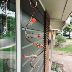 May include: A copper-colored hummingbird feeder with three glass vials and red caps. The feeder is designed with a spiral shape and is hanging outside a house. The feeder is attached to the window with suction cups.