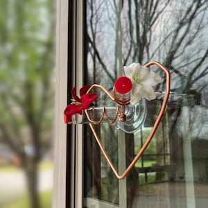 May include: A copper heart-shaped hummingbird feeder with a clear glass bottle and two red and white artificial flowers. The feeder is attached to a window with a suction cup.