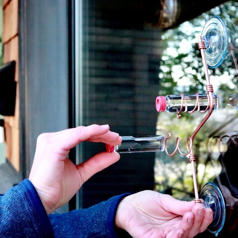 May include: A copper wire wall-mounted display rack featuring two clear glass vials with red caps. The rack is secured to a glass surface with two transparent suction cups. The wire is bent into spiral holders for the vials and straight sections.