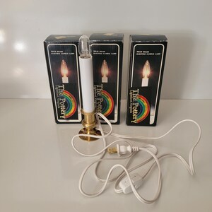 May include: Three electric candle lamps with white candle bodies, brass bases, and clear bulbs. The lamps are in black boxes with the text "The Pottery Lightfoot, Virginia" and a rainbow design. White cords and a switch are visible.